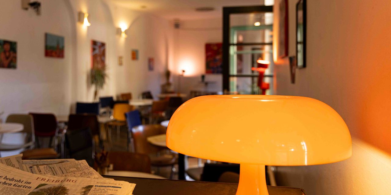 Eine leuchtende orangefarbene Retro-Tischlampe in Pilzform steht auf einem Tresen neben einer Tageszeitung.